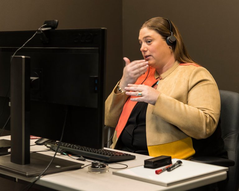 White woman with brown hair signing in front of a monitor