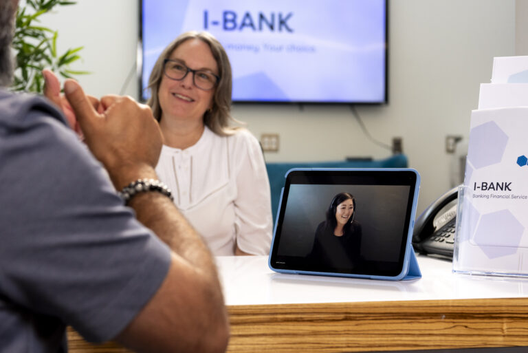 A white woman with glasses and a white shirt sits behind a desk at a bank. She is having a conversation with a Deaf customer using VRI On-Demand.