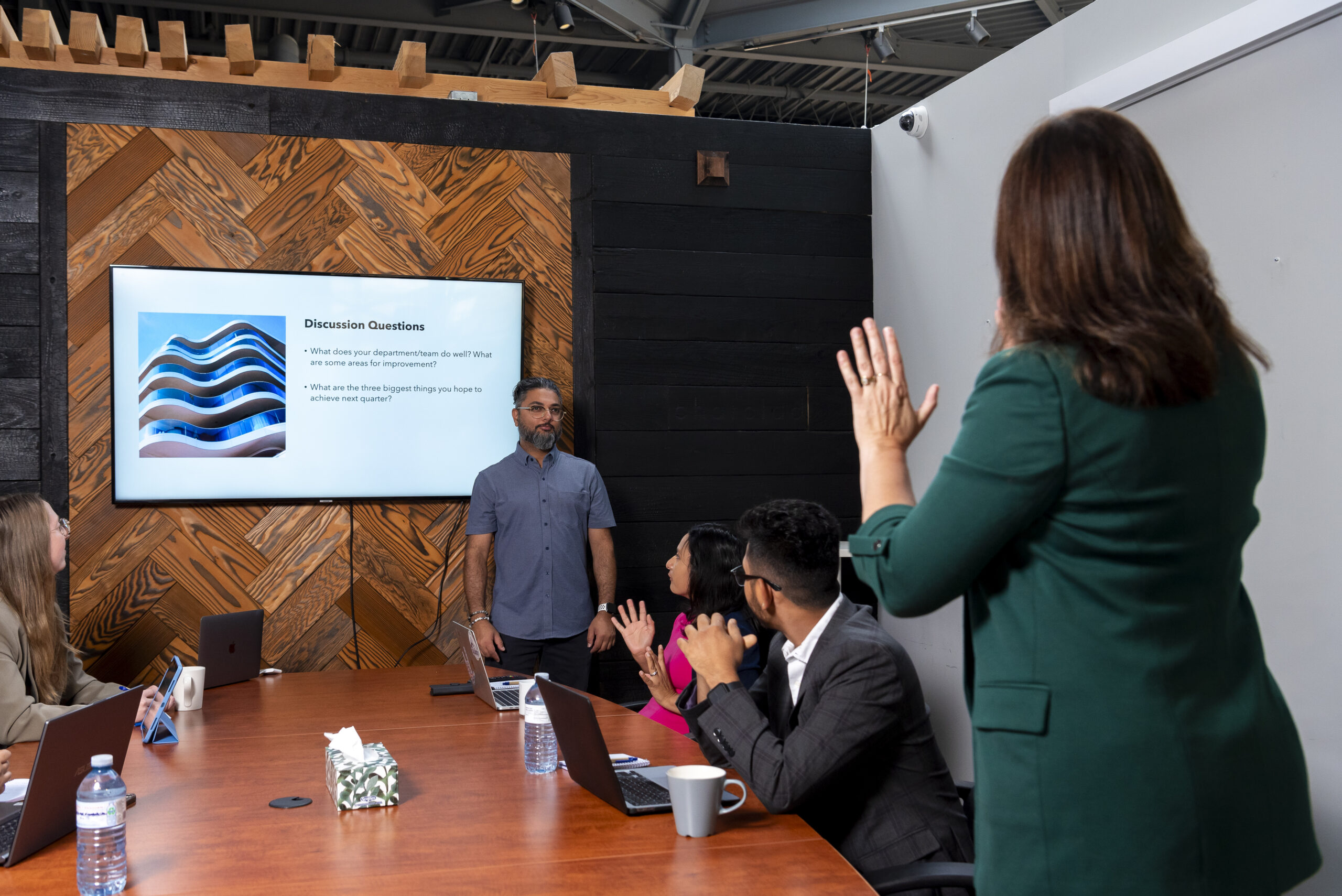 A man is standing at the front of a boardroom with a PowerPoint presentation behind him. Three people sit at the boardroom table all facing the man. A female interpreter is standing at the table interpreting the meeting.