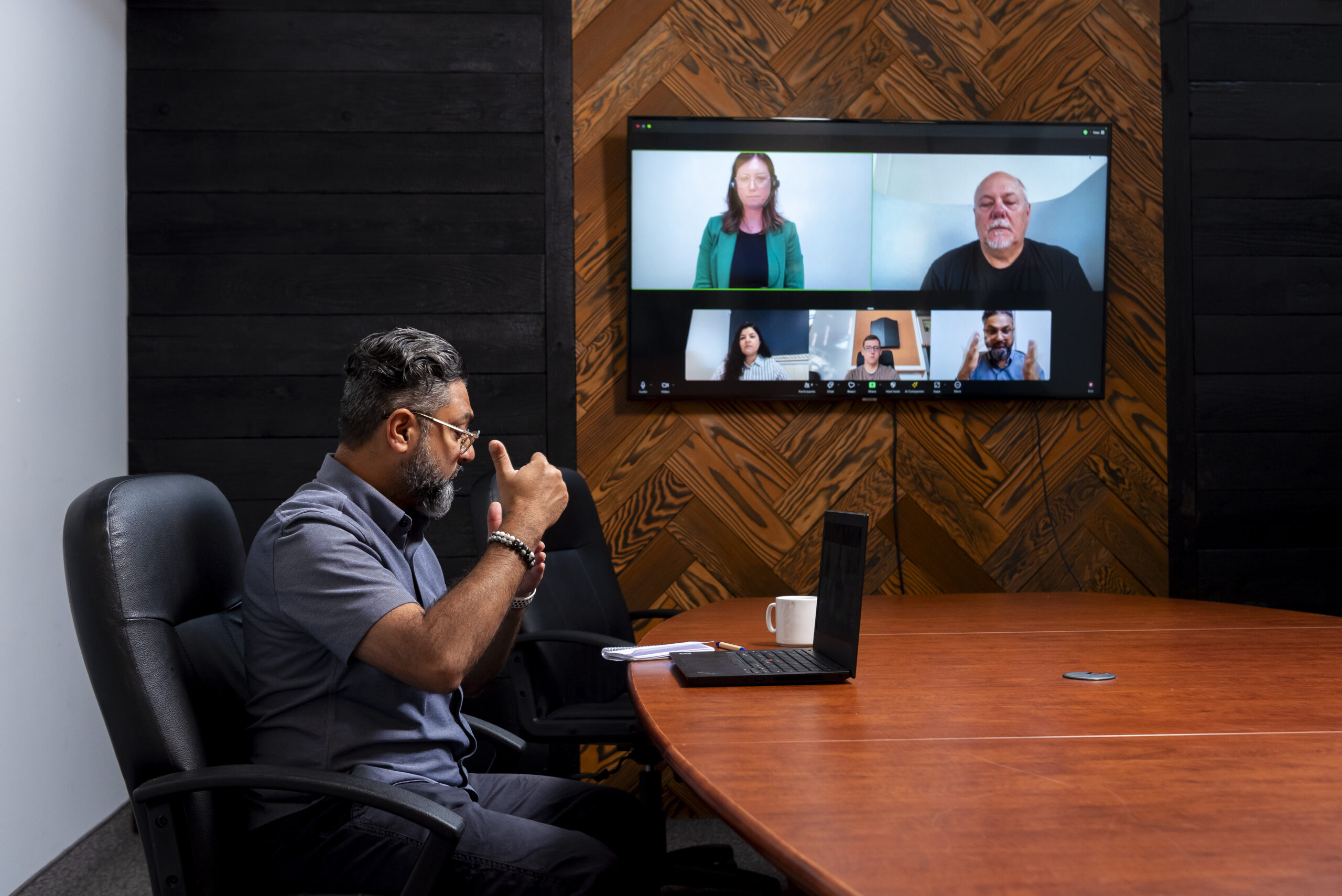 A man with gray hair and glasses signs with his team using Video Remote Interpreting (VRI) during a virtual meeting in a conference room. His laptop screen shows multiple participants, including an interpreter in a green blazer leading the discussion. A wooden panel wall with a large monitor forms the backdrop.