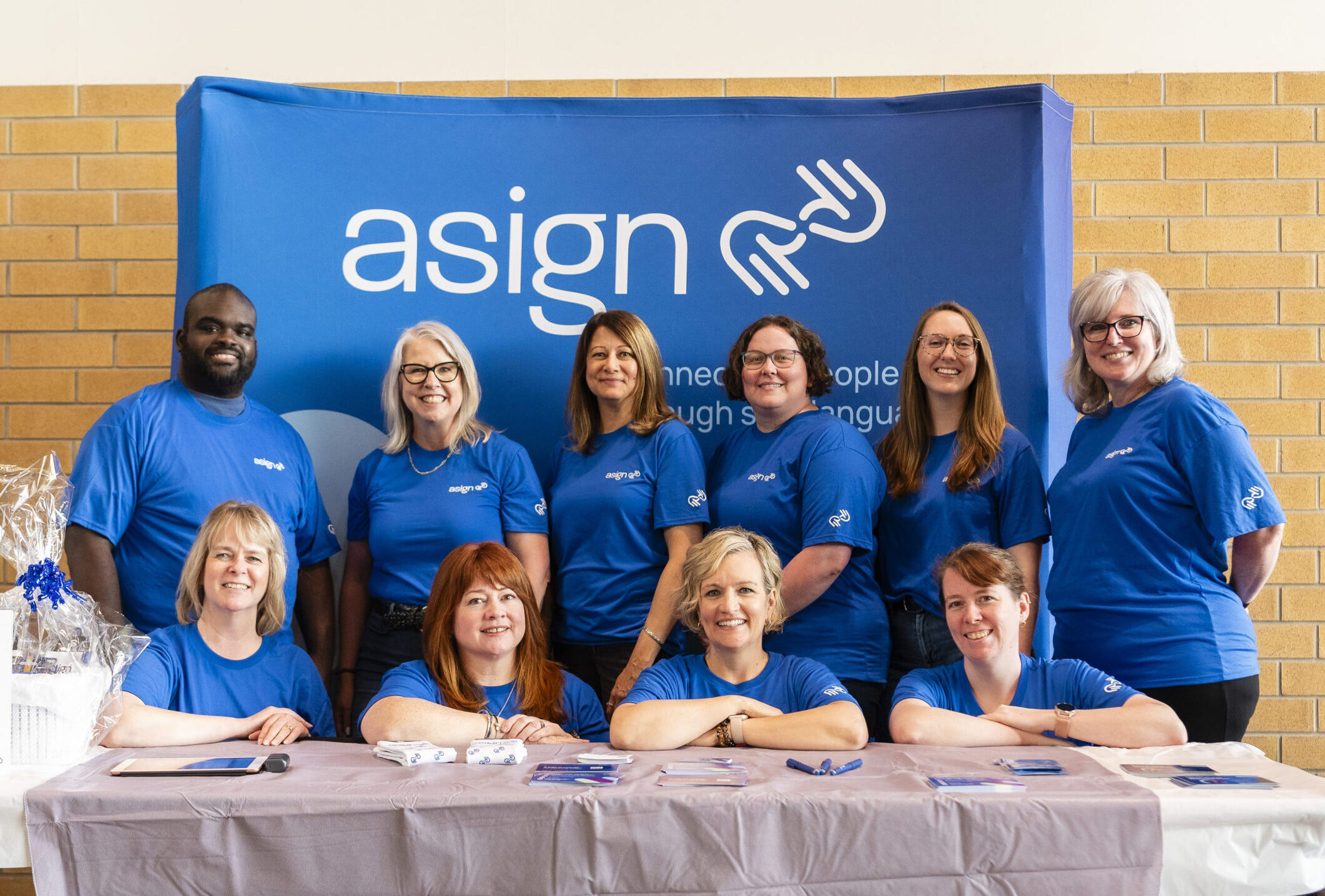 The Asign team smiling behind a table and in front of an Asign backdrop at Mayfest.
