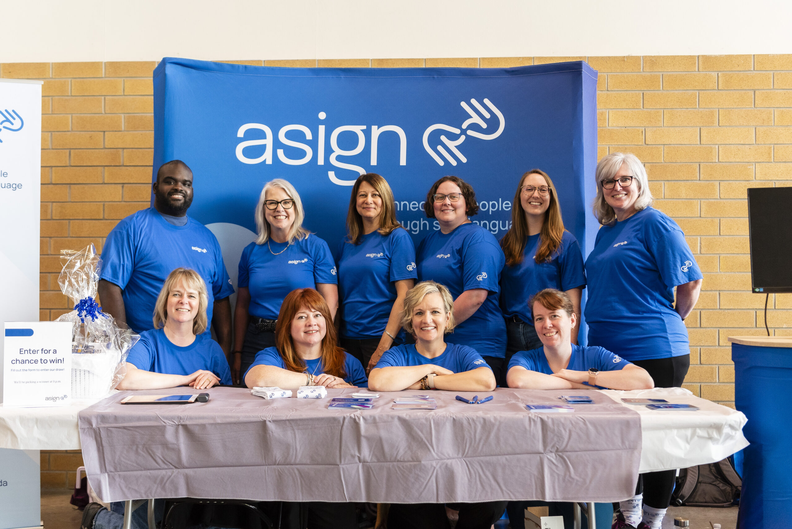 The Asign team smiling behind a table and in front of an Asign backdrop at Mayfest.