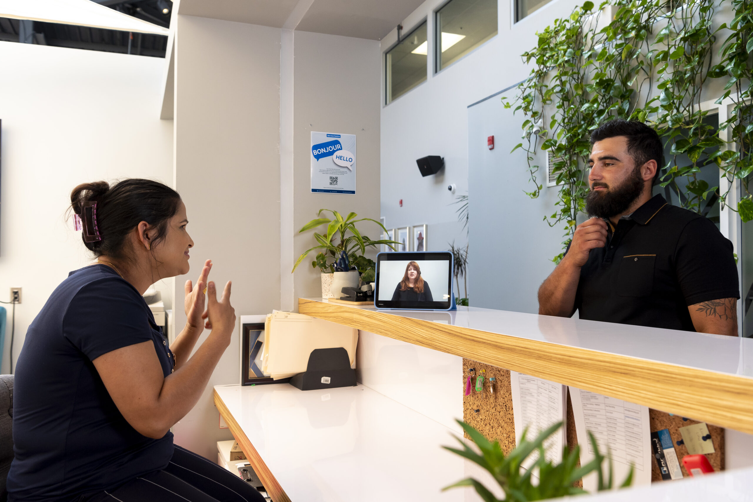 A woman with her hair pulled back in a bun sits behind a customer service counter. She is having a conversation with a bearded man standing in front of the counter using Video Remote Interpreting On-Demand.