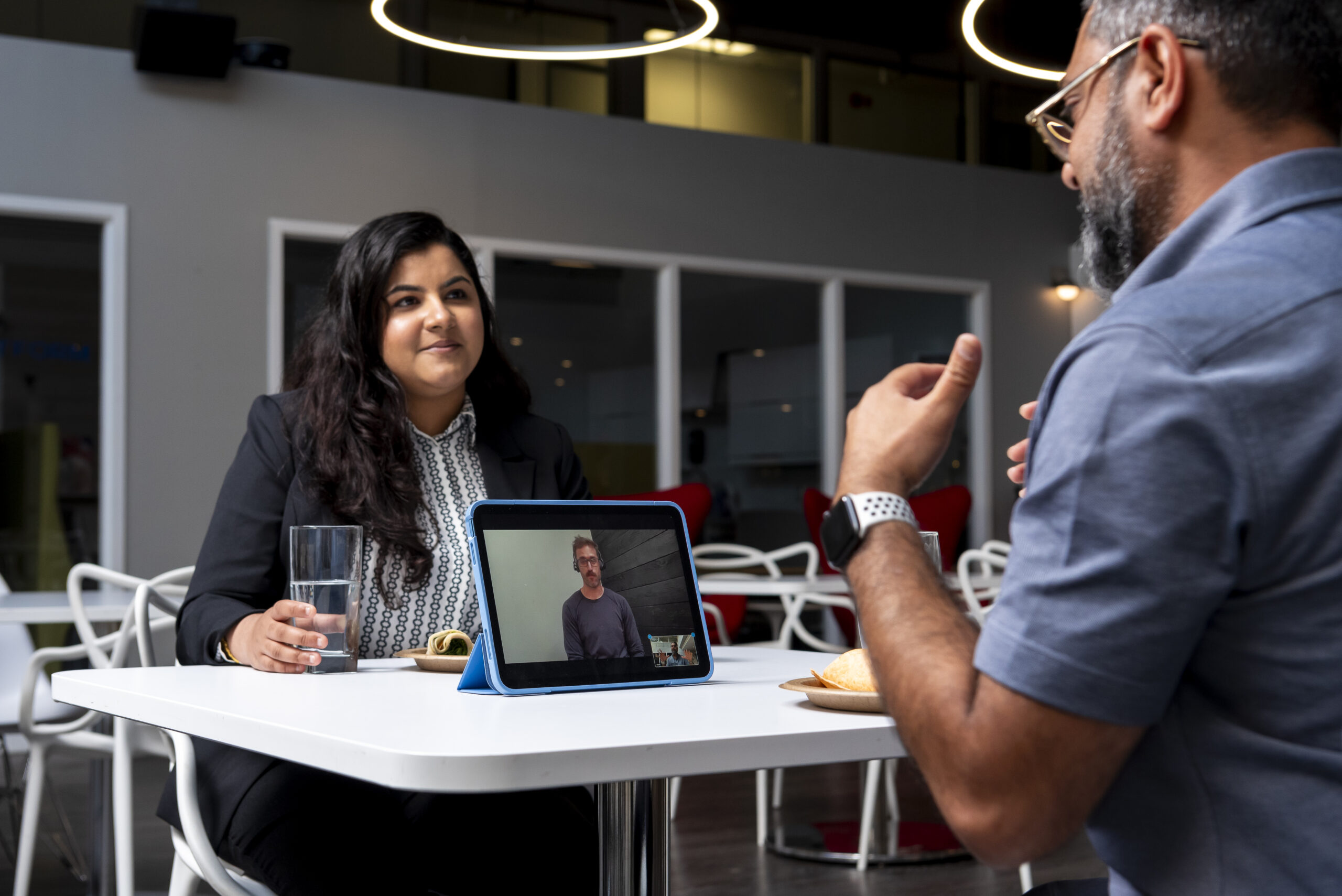 Partially in frame, a brown man with glasses and grey hair wearing a polo shirt is sitting at a white table behind an iPad. He is using VRI On-Demand to communicate with his colleague, a brown woman wearing professional attire, sitting on the opposite end of the table.