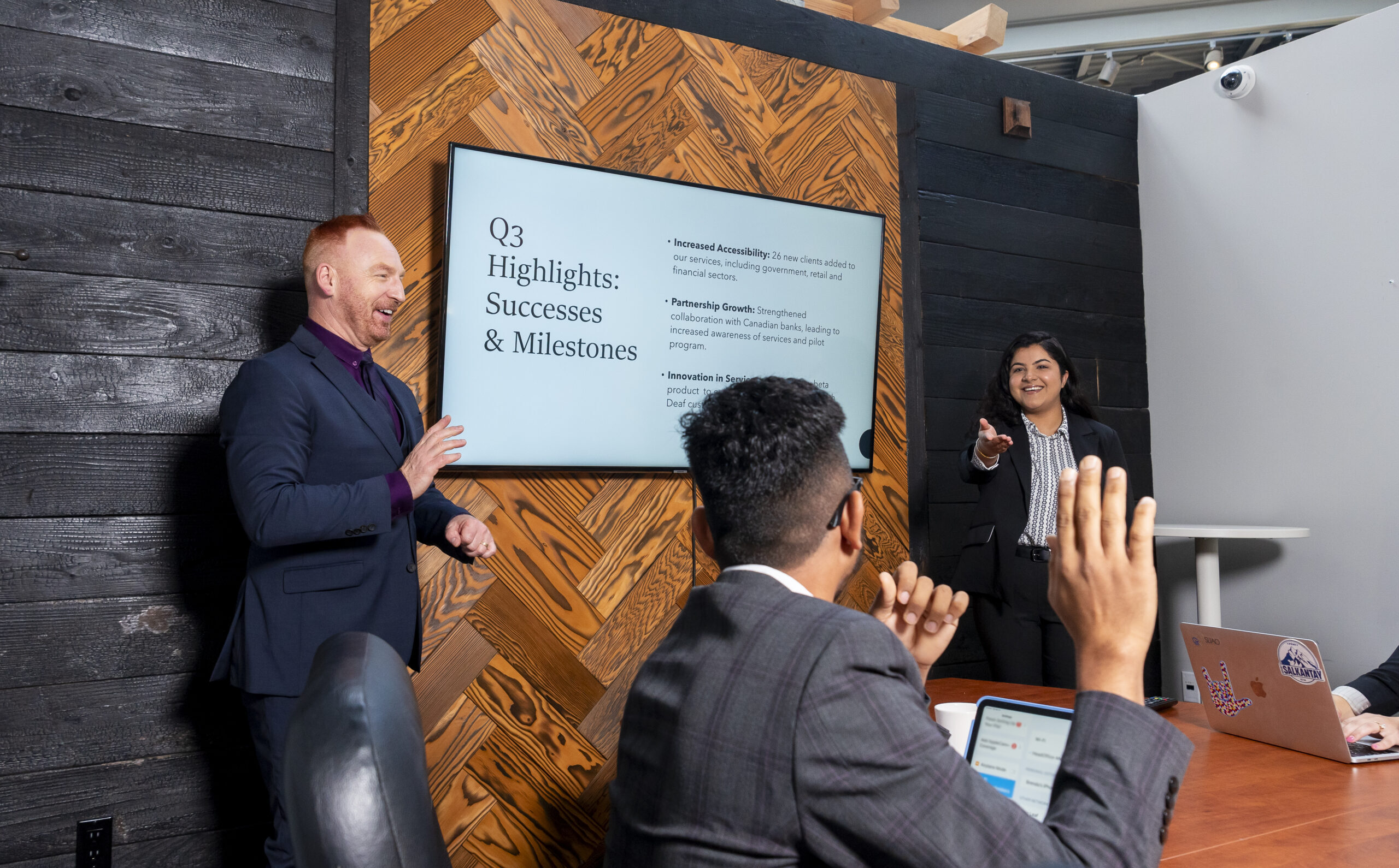 A woman is standing at the front of a boardroom with a PowerPoint presentation behind her. A man sitting at the boardroom table is raising his hand to ask a question. A male interpreter is standing at the front of the room interpreting the meeting.