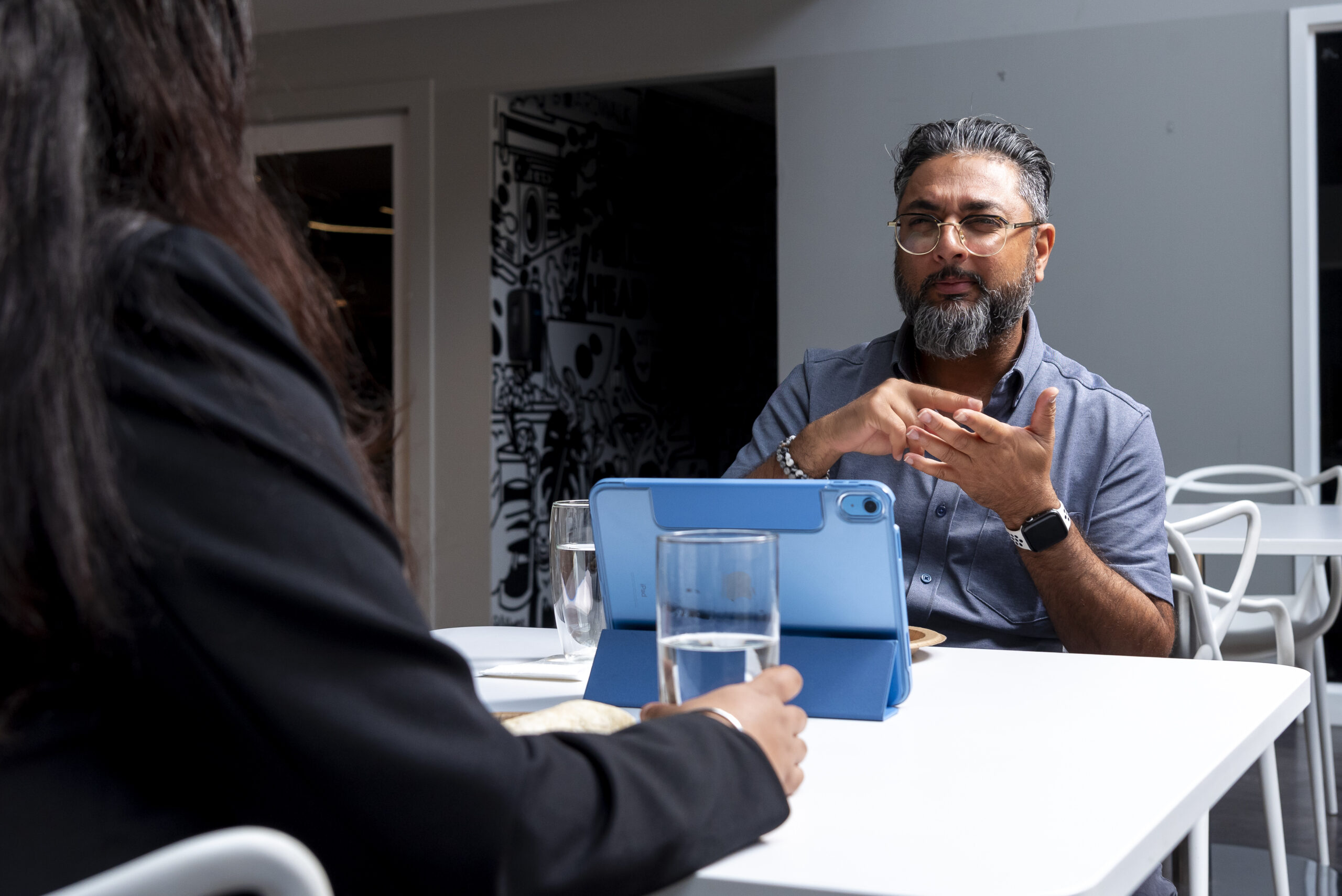 A brown man with glasses and grey hair wearing a polo shirt is sitting at a white table behind an iPad. He is using VRI On-Demand to communicate with his colleague.