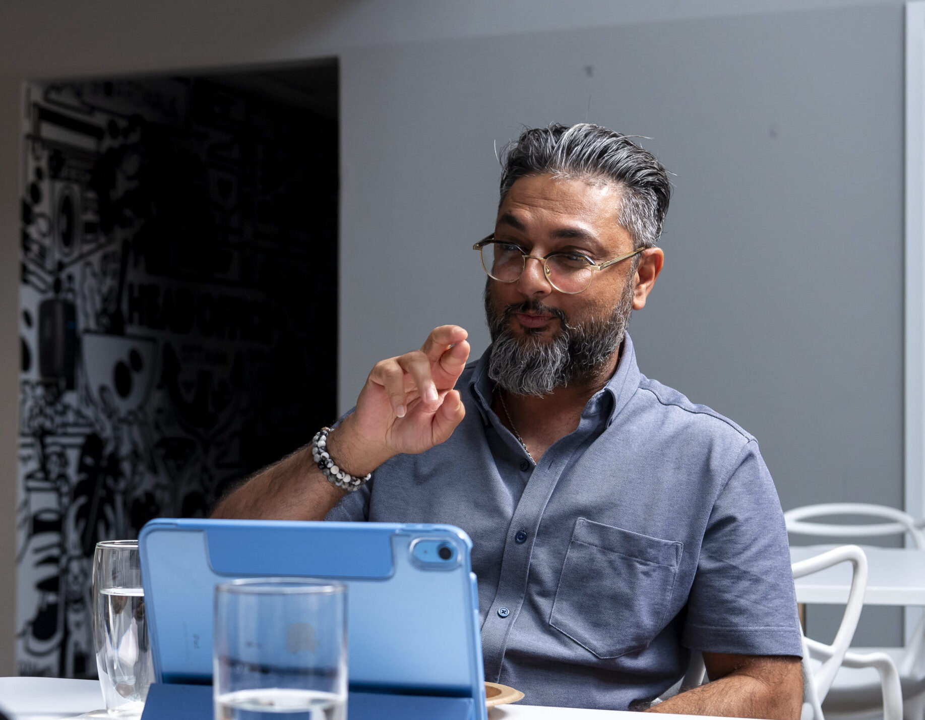 A brown man with glasses and grey hair wearing a polo shirt is sitting at a white table behind an iPad. He is using VRI On-Demand to communicate with his colleague.